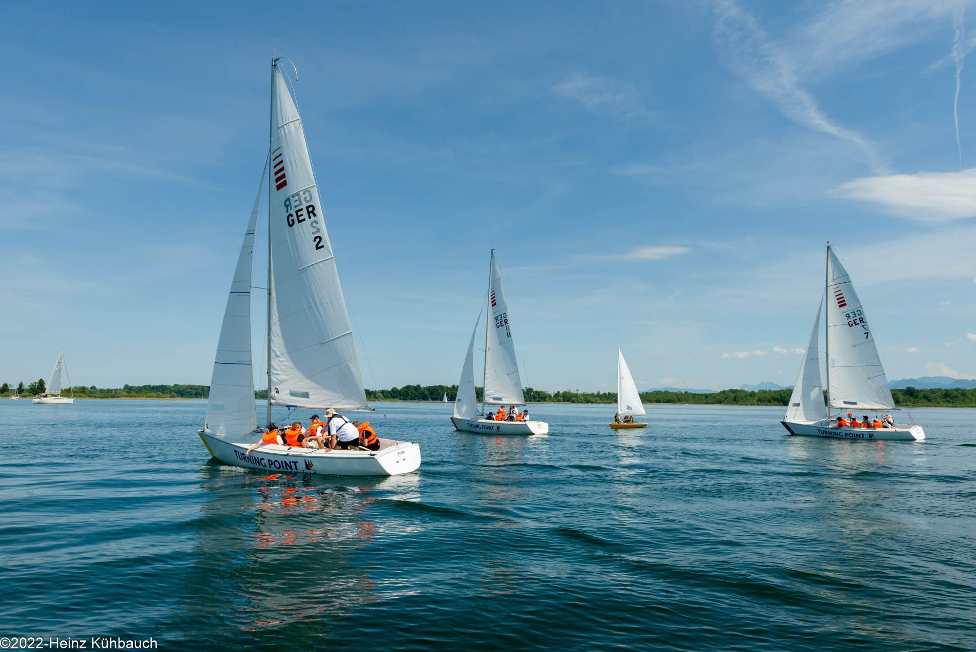 Es sind drei segelnde Boote der TURNING POINT Stiftung während eines Wendekurses auf leicht gekräuseltem Wasser zu sehen. Auf den Schiffen sind Kinder mit orangenen Schwimmwesten zu sehen.